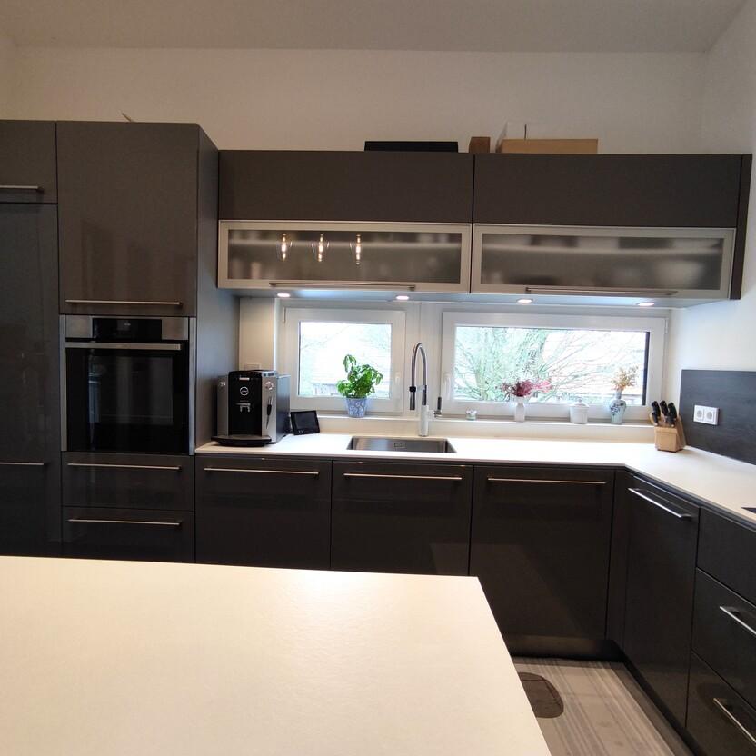 Moderne Küche nach Maß mit dunklen Hochglanzfronten, integrierten Geräten, weißen Arbeitsplatten und einer Fensterzeile für optimalen Lichteinfall und Stil.