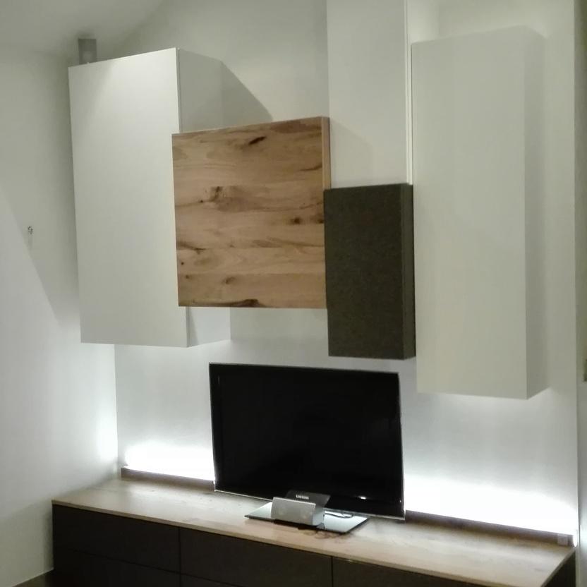 Moderne Wohnwand nach Maß mit hängenden Schränken, Holz- und Stoffelementen, sowie integriertem Beleuchtungssystem, ideal für eine stilvolle und funktionale Wohnzimmereinrichtung.