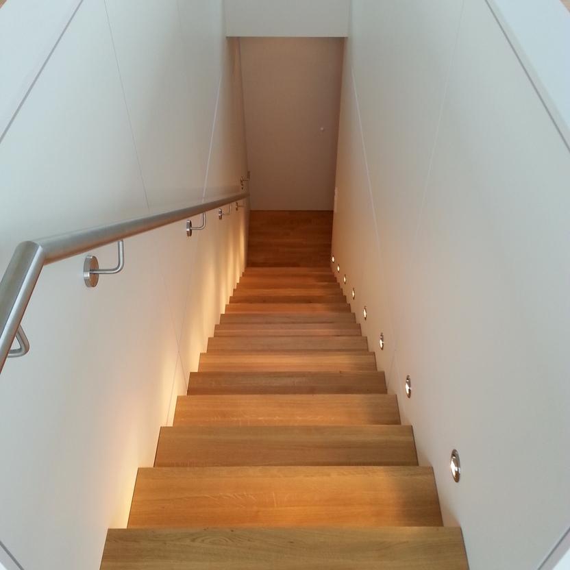 Maßgefertigte Treppe aus Holz mit integrierter LED-Wandbeleuchtung und Edelstahl-Handlauf, modern und minimalistisch gestaltet.