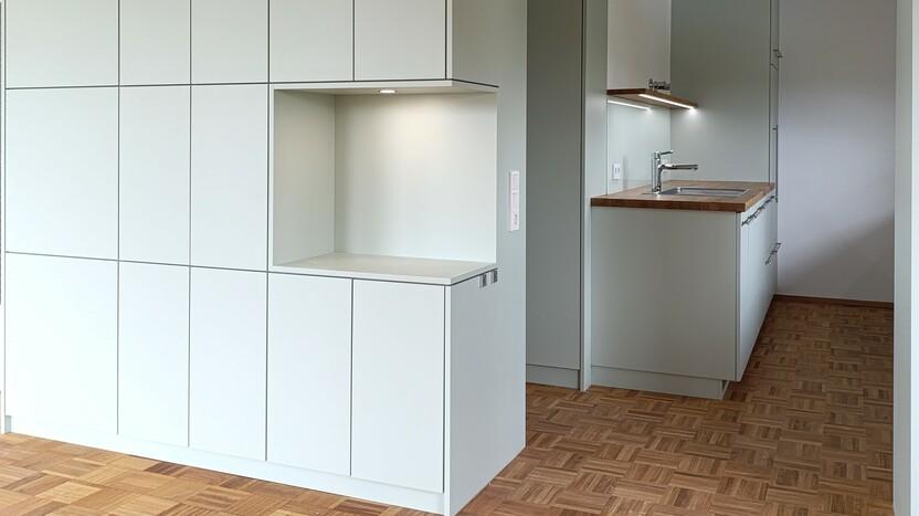 Schlichte Einbauküche mit grifflosen Fronten und integrierten Schränken, kombiniert mit Holzarbeitsflächen und moderner Beleuchtung für eine elegante und funktionale Küchengestaltung.