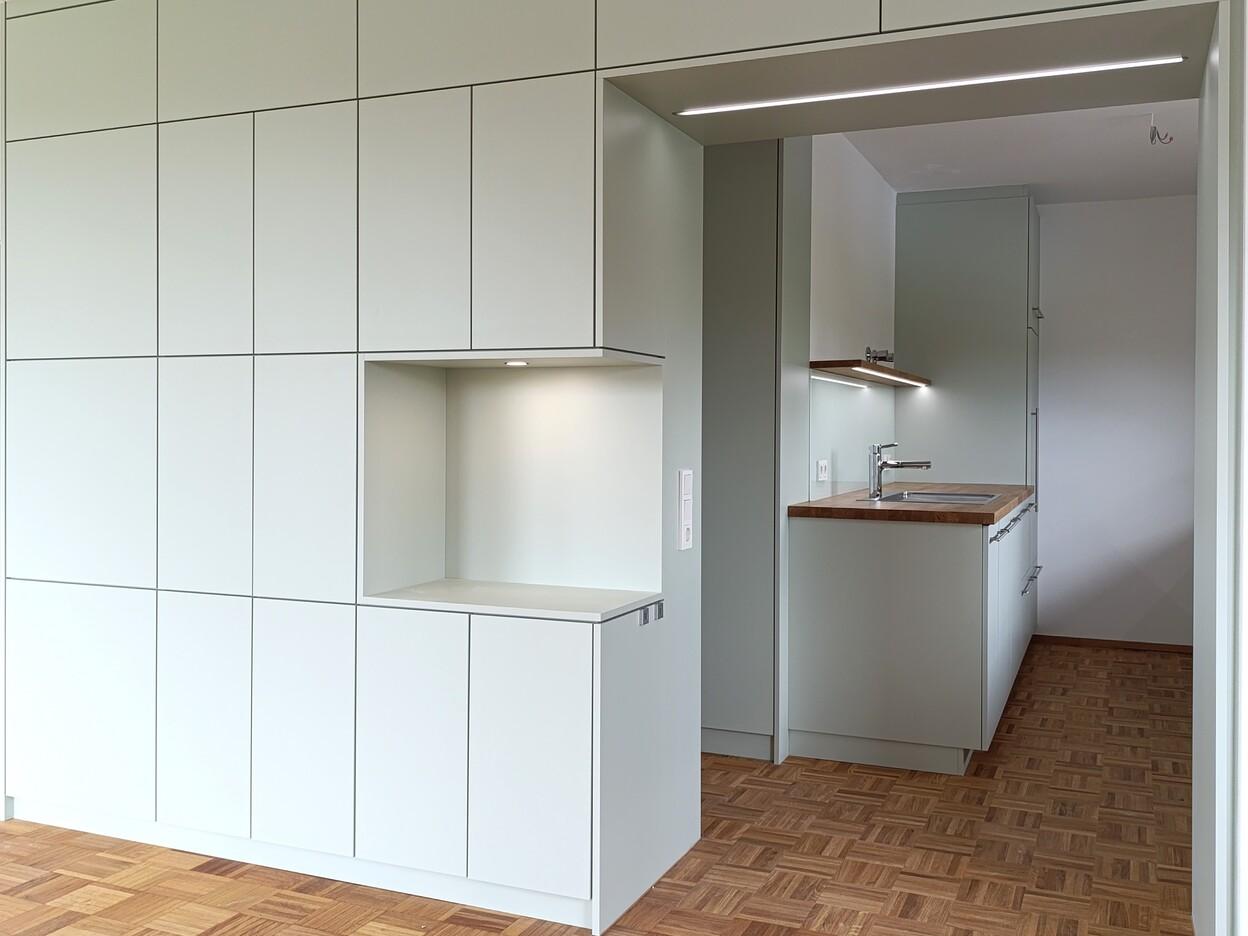 Schlichte Einbauküche mit grifflosen Fronten und integrierten Schränken, kombiniert mit Holzarbeitsflächen und moderner Beleuchtung für eine elegante und funktionale Küchengestaltung.